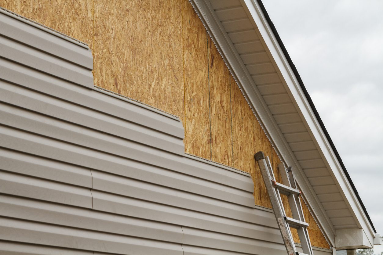 Tan vinyl lap siding being installed over OSB sheathing with a ladder against the eave